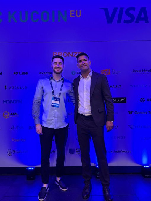 Two men pose together at the EBlockchain Conference in front of a step-and-repeat banner featuring "KUCOIN EU" and "VISA" branding alongside various sponsor logos like Lise, ARculus, and General Tokenization. One wears a light blue shirt with a lanyard, while the other is dressed in a dark suit, representing the Cryptic team's presence at Day 1 of the event as mentioned in the post.