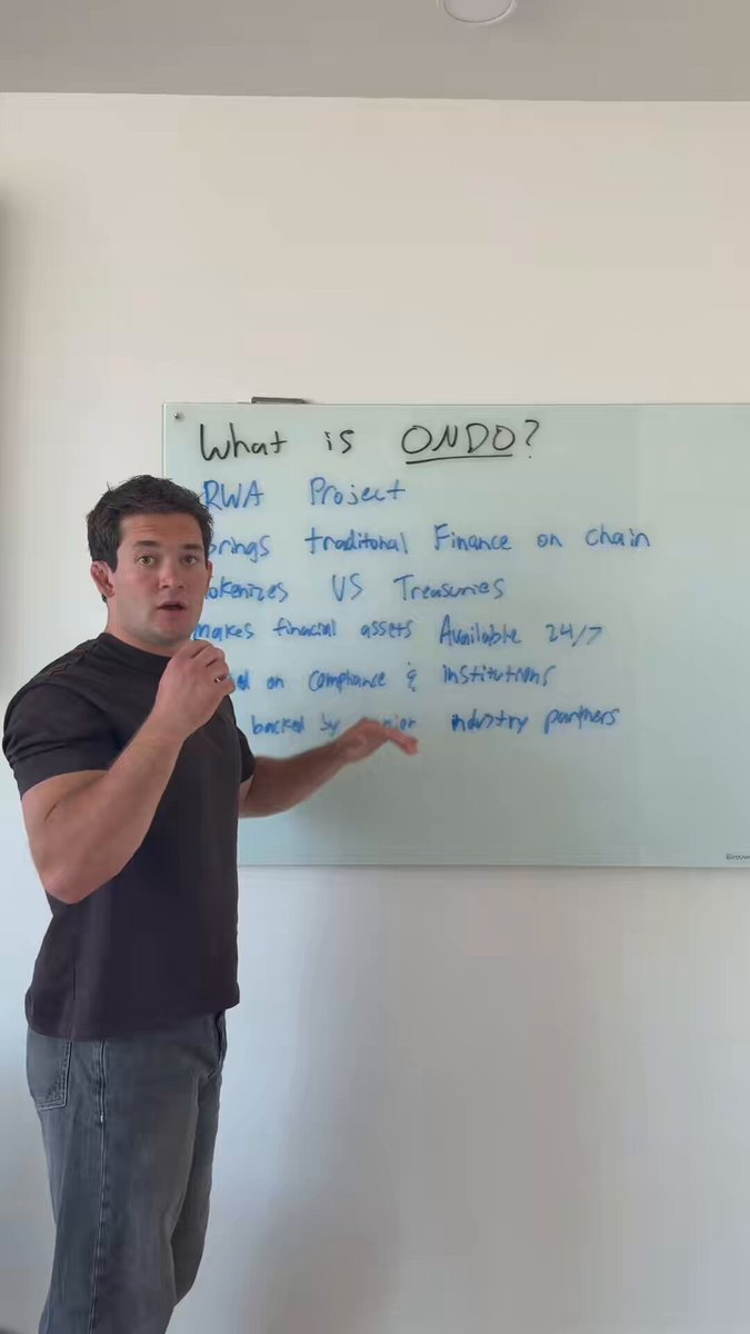 The image shows a man standing in front of a whiteboard, giving a presentation with his hands outstretched as if he's explaining something important or engaging an audience. The whiteboard has text written on it that reads "What is Ondo? #cryptotrading #blockchain #investment". The man appears to be discussing the topic of cryptocurrency and its potential impact on investing, possibly in relation to a project called Ondo.