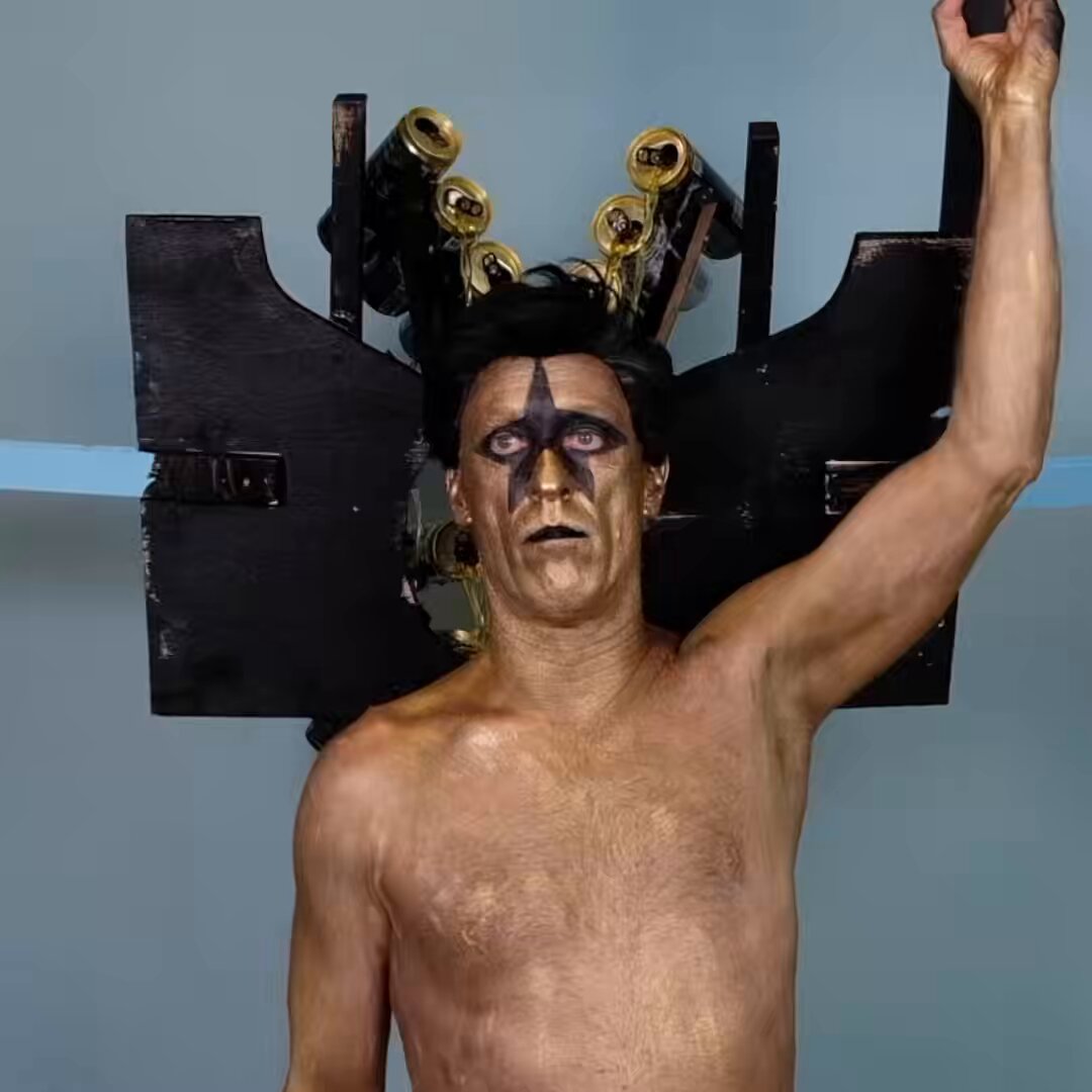 The image features a shirtless man with black hair, wearing a costume that includes a large number of gold cans attached to his head using a wooden frame. The man appears to be balding, and he has a serious expression on his face as he looks directly at the camera. The background is plain white, which contrasts with the colorful and eye-catching costume.