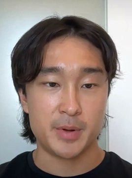 A close-up video call-style shot of a man with dark hair looking directly at the camera with his mouth slightly open.