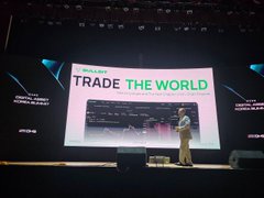 A presenter on a conference stage giving a talk titled 'TRADE THE WORLD' displayed on a large screen behind him.
