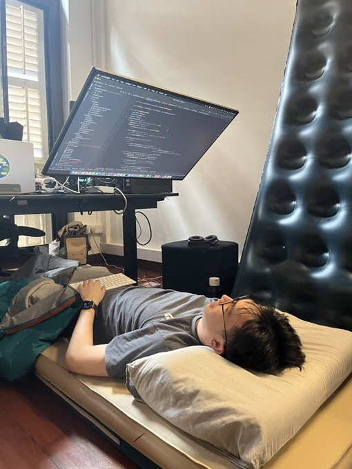 A person lies on a pillow and mattress on the floor, looking up at a large ultrawide monitor displaying lines of code, with VR goggles resting on an ottoman nearby. The setup captures the intensity of deep focus implied by "this is the level of locked in you need rn"—coding from a floor bed to maximize immersion and minimize distraction.