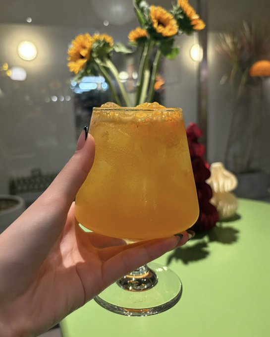 A hand holds up an orange-colored frozen cocktail in a stemmed hurricane glass, with sunflowers and a cozy ambient setting in the background. The refreshing drink pairs perfectly with the cheerful "good morning" vibe, suggesting a sunny start to the day.
