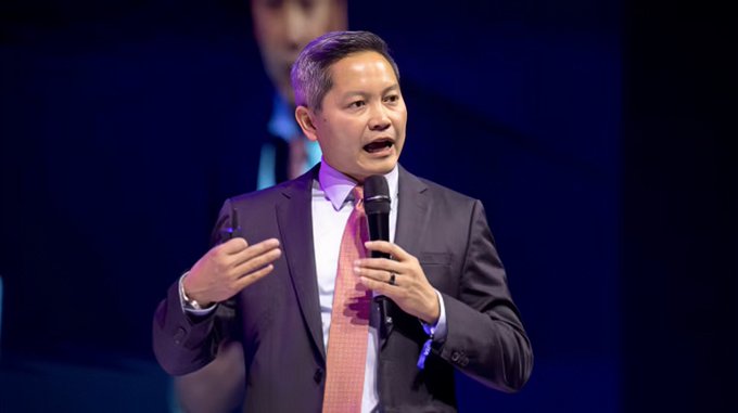 A man in a dark suit and pink tie speaking into a microphone on stage with a dark blue background, appearing to give a presentation or speech.