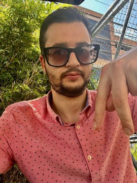 A man wearing sunglasses and a pink polka-dot shirt making a hand gesture towards the camera outdoors.