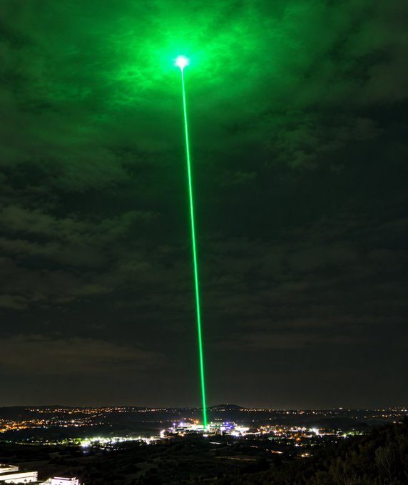 A brilliant green laser beam pierces the night sky, stretching from a glowing ground base up into the clouds, while the surrounding town twinkles with city lights below—visually capturing the "$RADRS to the moon" sentiment as the stock's trajectory aims skyward.