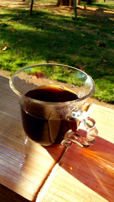 A clear glass mug filled with a dark liquid, possibly coffee or tea, sits on a wooden slatted table outdoors with green grass in the background.