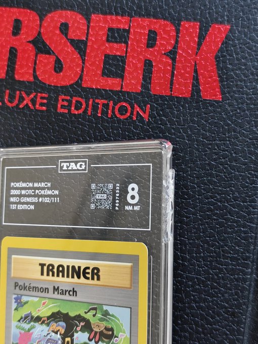 A graded Pokémon March Neo Genesis 1st Edition Trainer card (TAG 8 NM MT #102/111) sits in front of a Berserk Deluxe Edition book.