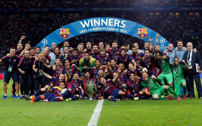A soccer team in blue and red striped jerseys celebrates as 'WINNERS UEFA CHAMPIONS LEAGUE 2015' behind a trophy display.