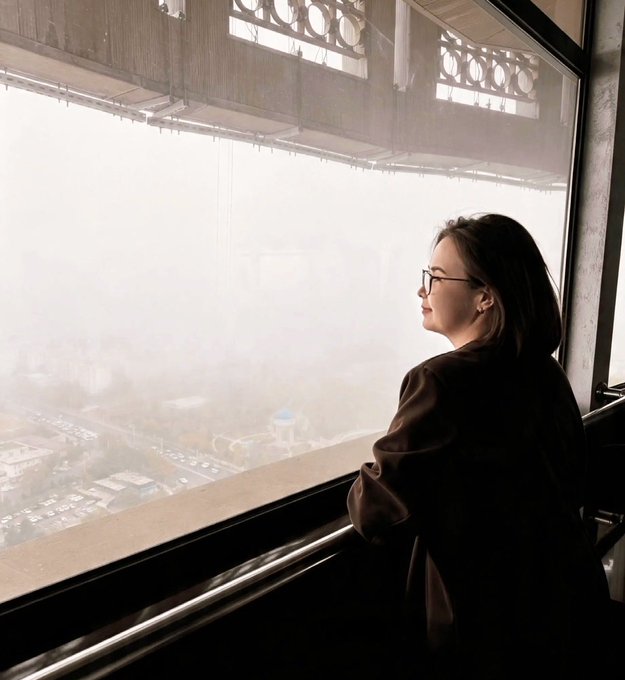 A woman wearing glasses and a dark jacket gazes out a large window at a foggy cityscape.