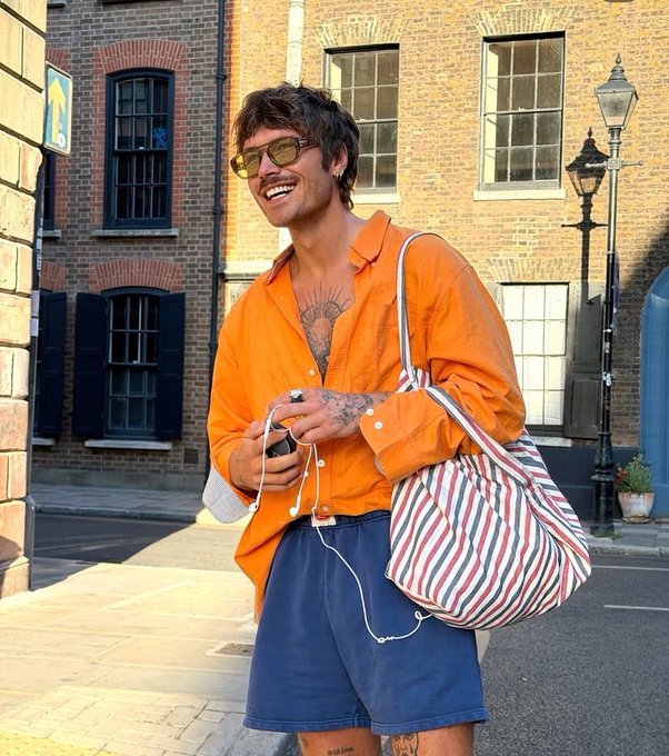 A man with tousled hair and sunglasses smiles while wearing an orange shirt and blue shorts, holding a striped tote bag on a brick-lined street.