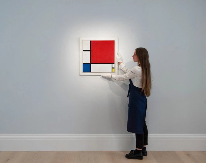The image features a woman wearing an apron, standing next to a painting of Piet Mondrian's "Composition with Red, Blue, Yellow, and White" (also known as the "Mondrian Square"). The painting is displayed on a white wall in a room. The woman appears to be examining or admiring the artwork, possibly considering it for sale or further study.