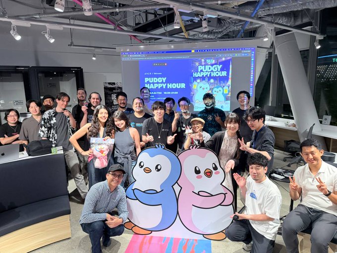 A group photo of a team posing in an office around two large cartoon penguin mascots (blue and pink), with a screen behind them displaying 'PUDGY HAPPY HOUR' event graphics.