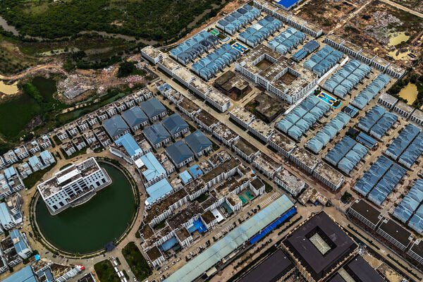 Aerial view of a large industrial complex or development area with numerous blue-roofed warehouses and factory buildings organized in grid patterns, surrounded by green vegetation and ponds.