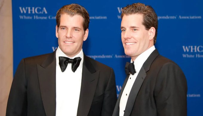 Twin men in matching black tuxedos with bow ties pose together at a White House Correspondents' Association (WHCA) event, standing in front of a blue backdrop with WHCA signage.