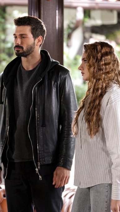 A bearded man in a black leather jacket standing next to a woman with long curly hair in a striped shirt.