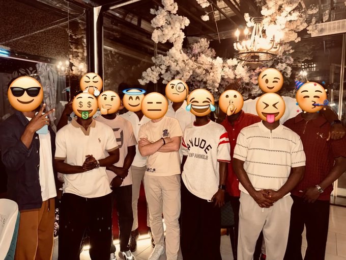 A group photo of several men standing together at an event with white floral decorations in the background, their faces obscured by various emojis.