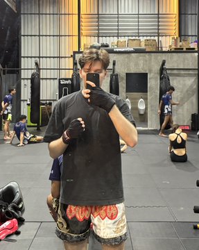 A man in a black t-shirt, boxing gloves, and flame-patterned shorts takes a mirror selfie at a gym.
