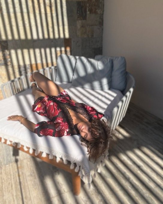 A woman with curly brown hair lies on a white daybed wearing a floral robe, bathed in striped shadows from a nearby window.