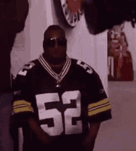 The image features a man wearing a black and yellow football jersey with the number 52, standing against a white wall. The background includes a clock and a picture frame, suggesting that this scene takes place at an event or gathering related to Bitcoin.