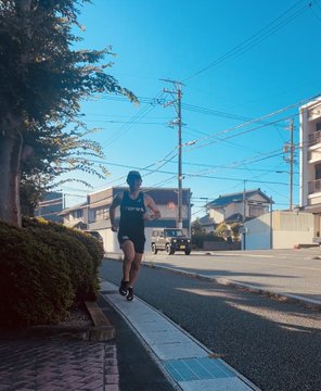 A slightly blurry photograph of a person jogging along a sidewalk in a residential neighborhood on a sunny day, with houses, parked cars, and utility poles visible in the background.