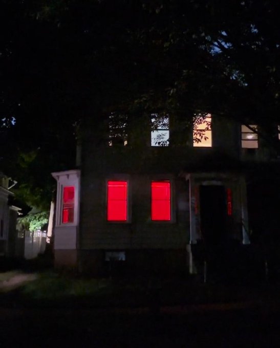 A nighttime photograph of a two-story house with its lower-level windows glowing bright red and upper windows illuminated with warm light, framed by dark trees.
