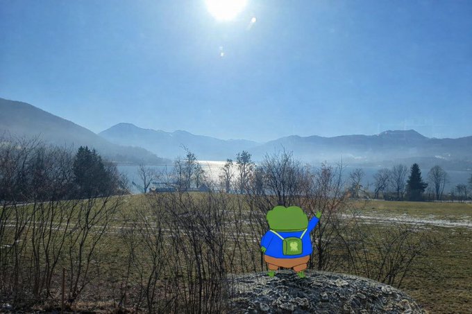 A small cartoon frog character with a blue backpack stands on a rock overlooking a misty lake and mountains in a scenic outdoor photograph.