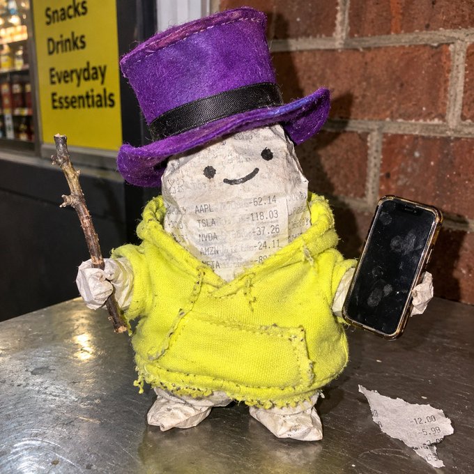 A whimsical paper-mâché figurine wearing a purple top hat and yellow robe holds a twig staff and a smartphone, with visible financial ticker data (AAPL, TSLA, NVDA, AMZN) printed on its body and nearby scraps of paper, playfully embodying the creator's journey from the $DEGEN experiment on the Base network to the official launch of $POOF on Robinhood Chain.