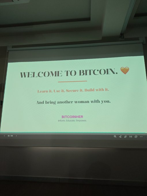 A projected presentation slide on a screen reading 'WELCOME TO BITCOIN.' with the text 'Learn it. Use it. Secure it. Build with it. And bring another woman with you.' under the heading 'BITCOINHER'.