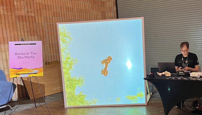 A man sits at a table with a laptop next to a large screen displaying the "Bones In The Sky Marfa" artwork and a corresponding poster on an easel, presented at the NFC Summit in Lisbon. The setup showcases the Art Blocks NFT piece as referenced in the accompanying post about "Bones In Marfa Balloons in Lisbon."