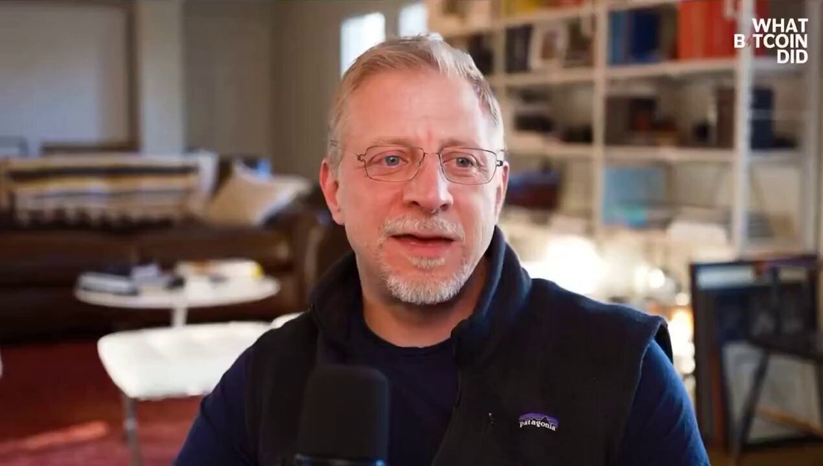 The image shows a man wearing glasses, sitting inside a room with bookshelves around him, which appears to be an office or library setting. The main focus is on the man, who has a microphone positioned over his head as if he is giving a speech or presentation. The title of the photo reads "James Lavish Says Bitcoin Is Going Through a Information Arbitrage and I Think That Bitcoin is gonna trade above $1,000,000 in early 1903s." This title suggests that James Lavish, an expert in Bitcoin technology, is discussing the impact of Bitcoin's information arbitrage potential and its projected growth to be above $1,000,000 in early 1903.