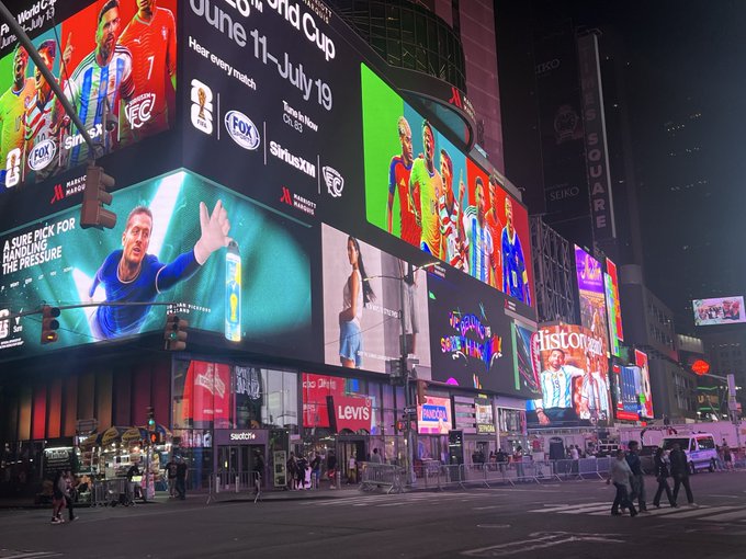 The image shows Times Square at night, illuminated by bright digital billboards advertising the 2018 FIFA World Cup, FOX Sports, and SiriusXM, along with other advertisements. The post references NFTs at NFT_NYC 2026, though the image itself appears to be from the 2018 World Cup event and doesn't visibly feature any NFT-related content.