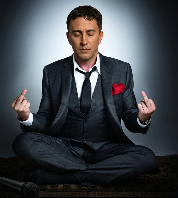 A man in a charcoal three-piece suit with a red pocket square sits cross-legged in a meditation pose with his eyes closed, while a microphone lies discarded on the ground beside him. The image visually reinforces the "$hotemin still HOT" caption by juxtaposing serene meditation with stand-up comedian attire, suggesting a performer finding inner calm amid the comedy spotlight.