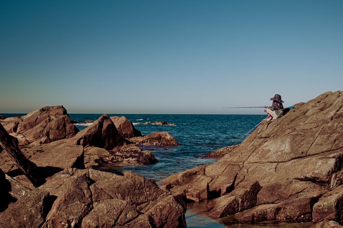 A person sits on rugged brown coastal rocks casting a fishing line into the deep blue ocean under a clear sky.