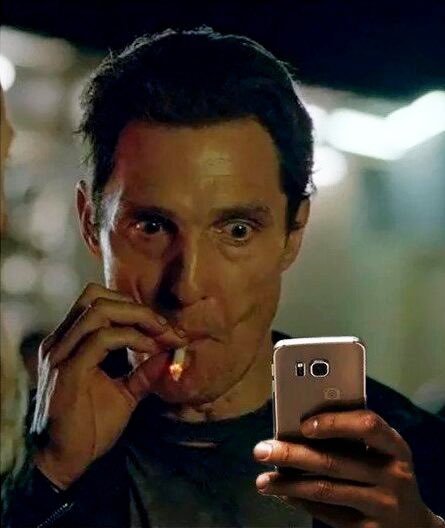 A close-up shot of a wide-eyed man smoking a cigarette while intently looking at a rose gold smartphone at night.