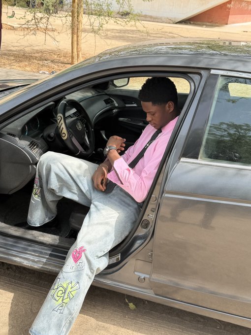 A young man wearing a pink shirt and light-wash jeans with graffiti-style designs sits in the driver's seat of a car, casually checking his watch. The image conveys a relaxed, on-trend vibe that complements the "GM Web3 We are winning today" post, symbolizing a stylish and confident approach to the Web3 lifestyle.