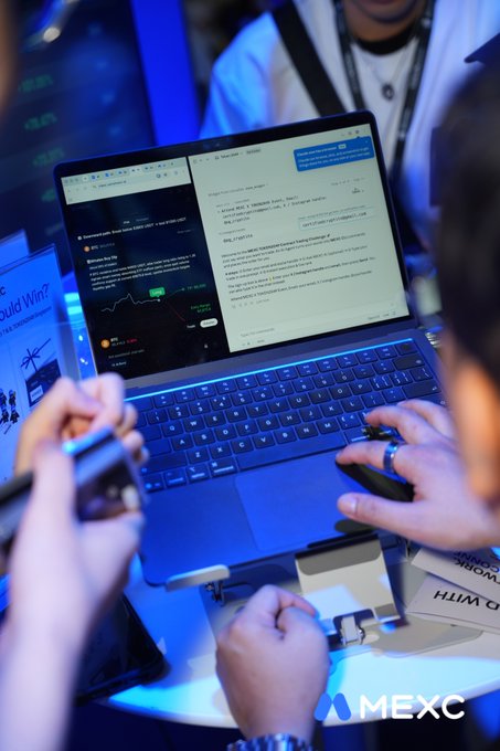 Two participants compete side-by-side at the MEXC CLI Trading Challenge, with one laptop displaying a real-time BTC/USDT price chart while the other shows a leaderboard of top traders, including names like "Abhi Z" and "Brainy Boy." The MEXC-branded setup, blue ambient lighting, and "Should Win?" signage reflect the competitive event atmosphere highlighted in the post's "TradingTakesTwo #MEXCEventRocks" promotion.