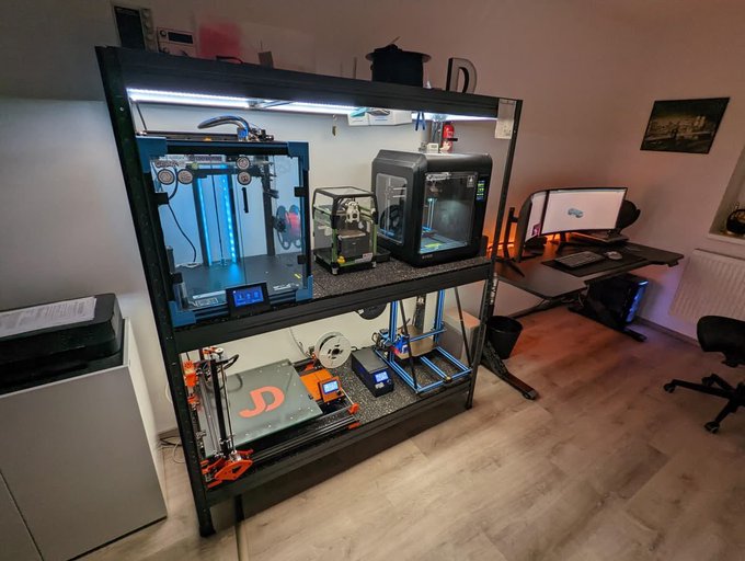 The image shows a well-equipped 3D printing workspace featuring a multi-level shelving unit housing various 3D printers, including models with visible "BIBO" branding and what appears to be a "JD" logo, alongside a desktop computer setup with dual monitors. This relates to the post's discussion of Web3 and NucleusCodes, as 3D printing represents tangible, productive value creation—the kind of meaningful output that contrasts with the empty engagement metrics the post criticizes.
