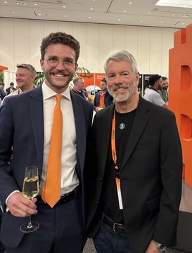 Two men in suits pose for a photo at an event, with the man on the left wearing an orange tie and holding a glass of champagne.