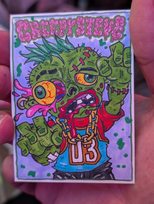 A person holding up a small, colorful illustrated sticker or card titled 'GREASYSLIM' featuring a grotesque green zombie-like character wearing a '03' basketball jersey.