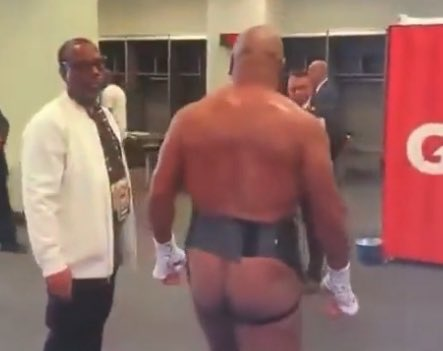 The image shows a muscular, nearly naked bodybuilder standing backstage, partially exposing himself, while a man in a white shirt and tie looks on in a locker room setting. This appears to be a humorous meme referencing Bitcoin enthusiasts anxiously watching for the cryptocurrency to dip below $77K, with the bodybuilder's exposed state symbolizing their "bare" vulnerability and eagerness for a price drop to buy in.