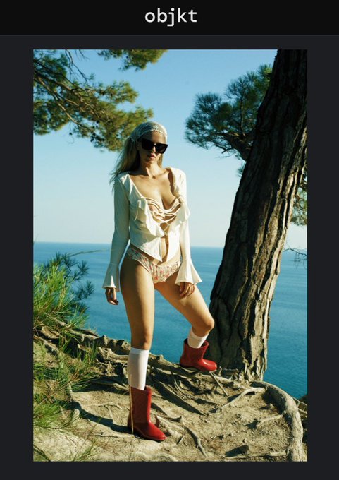 A fashion photograph shows a woman wearing sunglasses, a white corset top, lace shorts, and red boots standing by a pine tree on a cliff overlooking the ocean, with 'objkt' at the top.