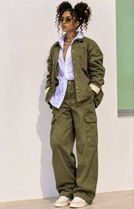A woman models a streetwear outfit consisting of an oversized olive green jacket, matching cargo pants, a light blue button-down shirt, and beige sneakers.
