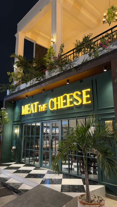 An illuminated storefront at night displays a bright yellow neon sign reading 'MEAT THE CHEESE' above green-framed glass doors, with a checkered marble entrance and potted palm plant.