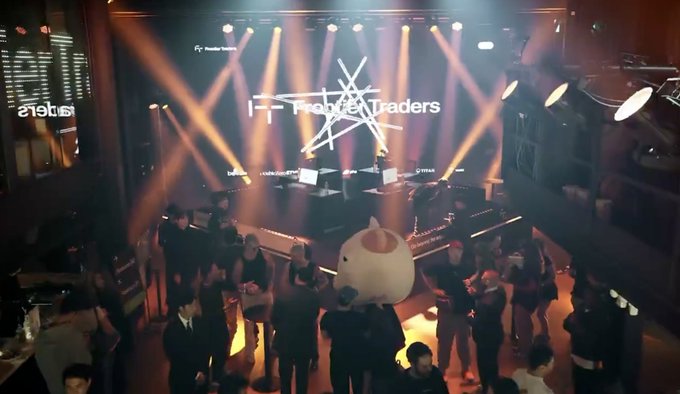 Photo of a crowded event venue with stage lighting and a screen displaying 'FractureTraders', with a large mascot character in the center of the crowd.