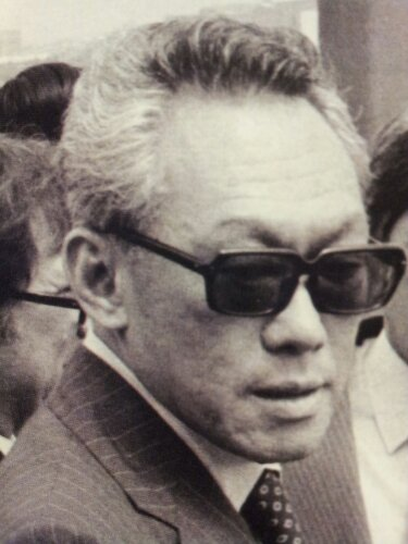 A black-and-white photograph of an older man wearing thick, dark-rimmed glasses and a suit with a patterned tie.
