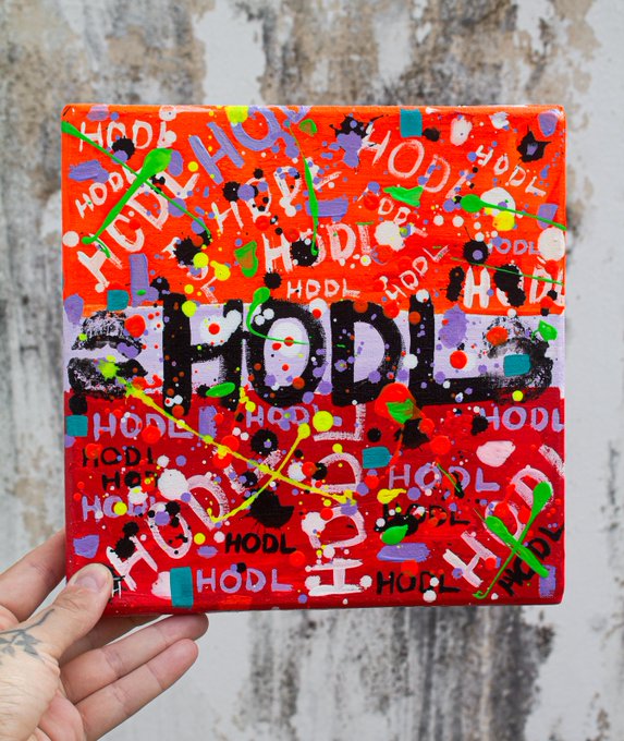 A hand holds a small square canvas artwork with a vibrant red background covered in multicolored paint splatters and the word 'HODL' repeated throughout, with a large black 'HODL' in the center.