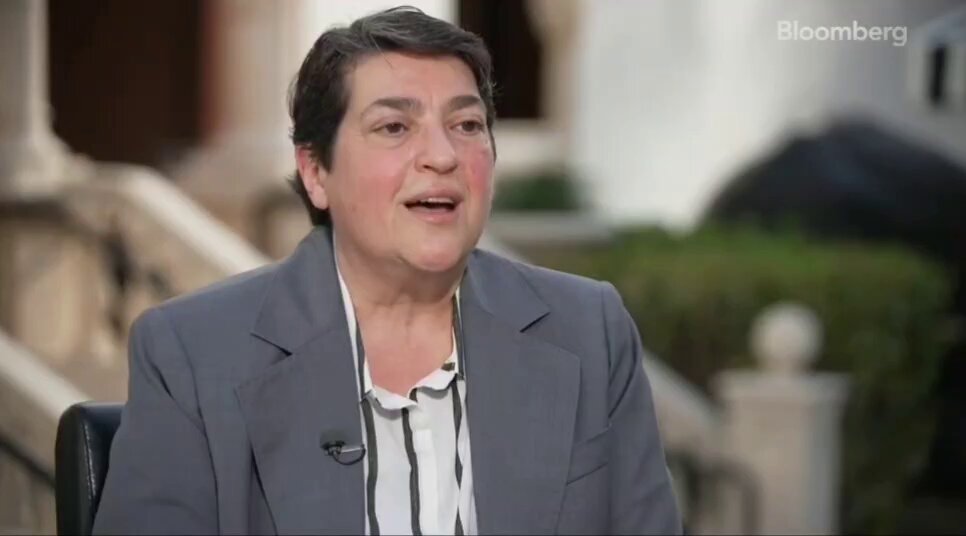 The image shows a woman wearing a gray suit jacket, sitting down with her hands clasped together. She appears to be engaged in conversation or giving an interview, as indicated by the presence of a microphone behind her. The background features a building and some bushes, suggesting that she might be in an urban setting. The post also includes text about the date "July-October 2026 #RipplePrime #Stellar #Canton".