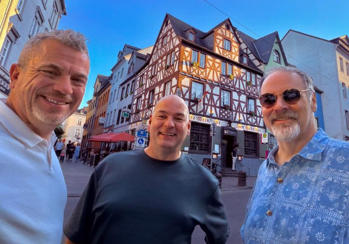 A selfie of three smiling men standing in front of traditional half-timbered German buildings on a street corner.