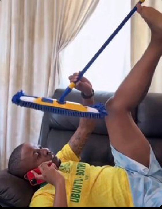 A humorous scene of a man in a yellow "UBUNTU BOTM" shirt lying upside down on a couch, talking on a red phone with his foot, while holding a blue and yellow mop against his other foot.